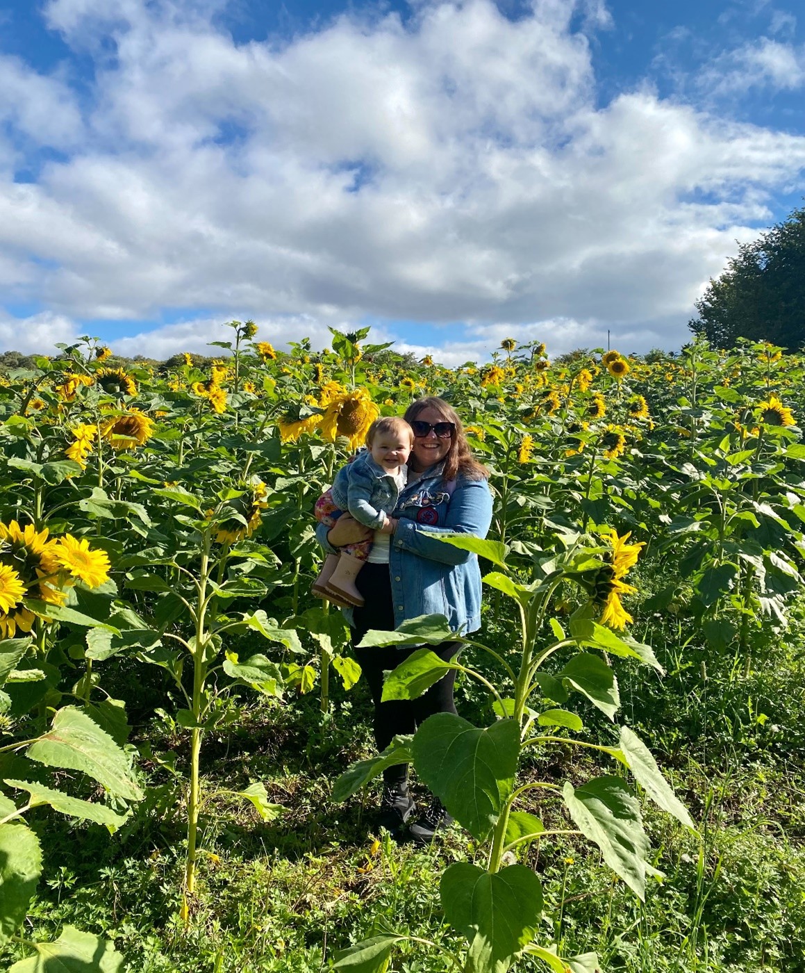 Photograph of Leigh Miele, Training Co-Ordinator for Employability at UHI Moray, standing in a sunflower field and holding her daughter