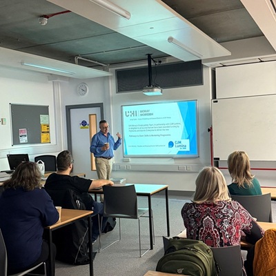 Photo of a male dressed in a blue shirt and tan trousers presenting to a room of people in UHI Moray