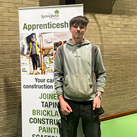 Photo of Ben next to a Springfield pop-up banner at an event. The background is a brick wall and green floor.