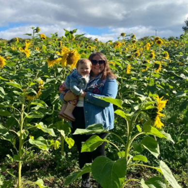 Photograph of Leigh Miele, Training Co-Ordinator for Employability at UHI Moray, standing in a sunflower field and holding her daughter Photograph of Leigh Miele, Training Co-Ordinator for Employability at UHI Moray, standing in a sunflower field and holding her daughter