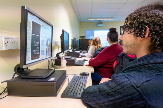 Photo of a student reading a document on a computer screen, in the background students are looking at a presentation Photo of a student reading a document on a computer screen, in the background students are looking at a presentation