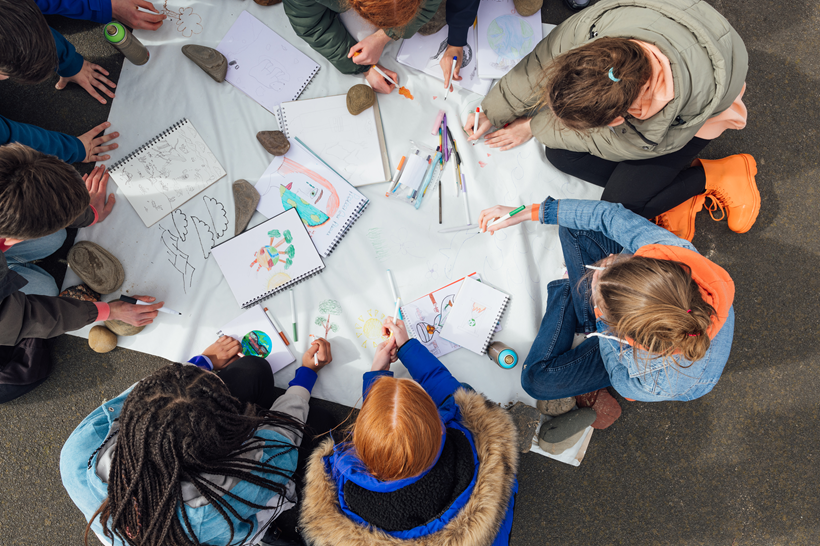 Top down photo of children around a big piece of paper doing a colouring activity outdoors Top down photo of children around a big piece of paper doing a colouring activity outdoors