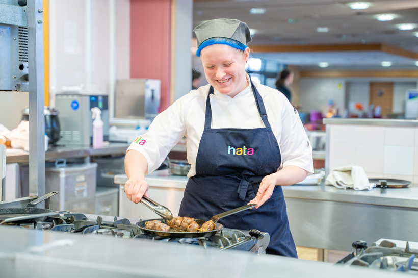 Photo of a female chef in a kitchen cooking. In the background you can see waiting staff guiding guests to their seats in the restaurant Photo of a female chef in a kitchen cooking. In the background you can see waiting staff guiding guests to their seats in the restaurant