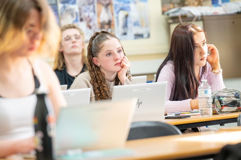 A photo of classroom environment with a female student in focus looking forward with her head in her hands. A photo of classroom environment with a female student in focus looking forward with her head in her hands.