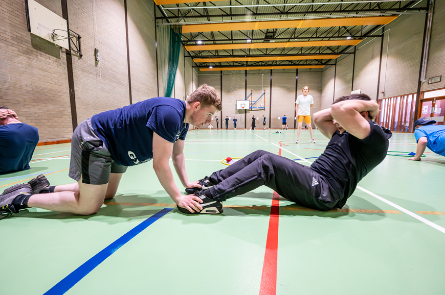 Photo of two male students doing an exercise in the games hall. One student is practising being a personal trainer to the other Photo of two male students doing an exercise in the games hall. One student is practising being a personal trainer to the other